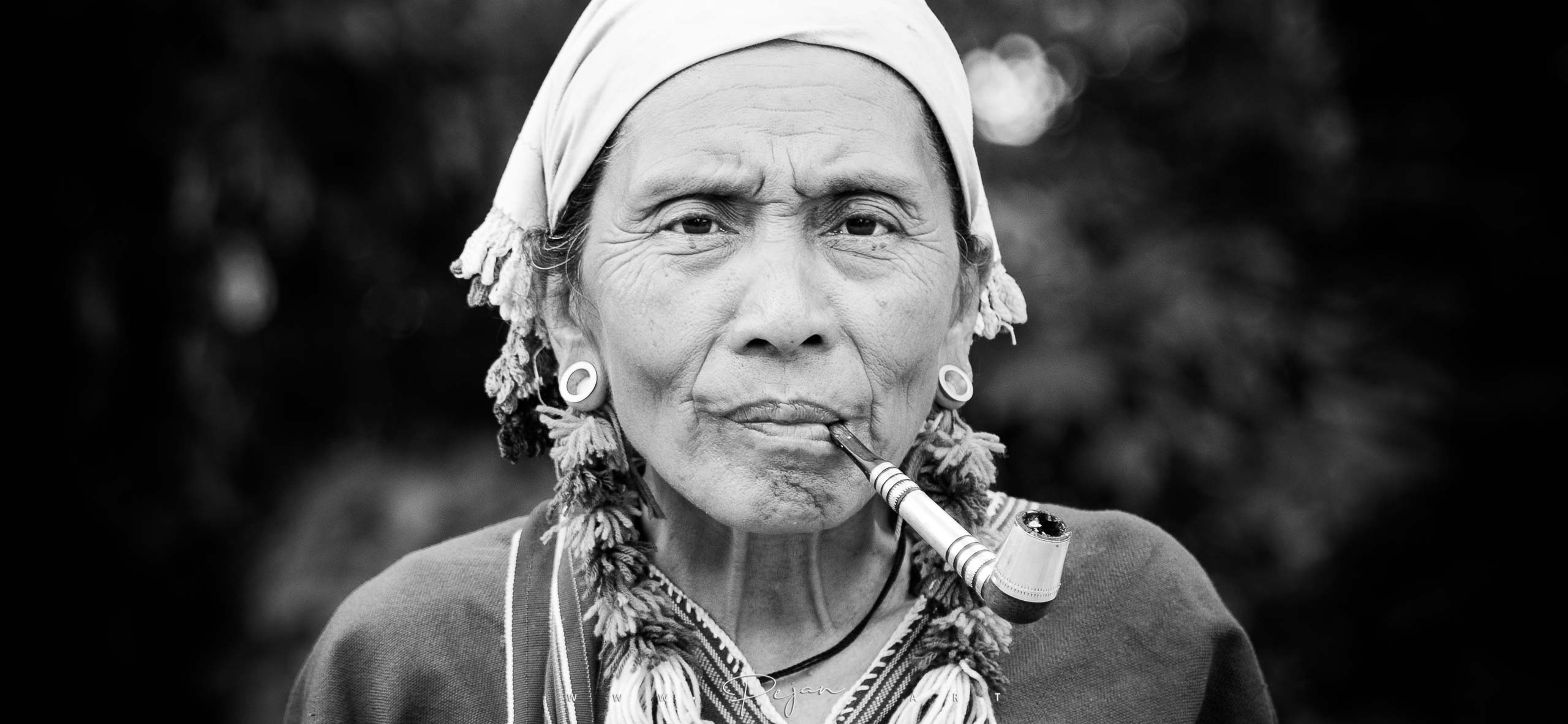 Monochrome portrait of a woman from the Karen ethnic group encountered in the mountains in Thailand. She wears traditional clothing and jewelry and smokes a large pipe. She looks at the camera.