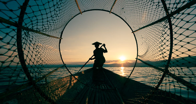 Carnet de voyage en Birmanie - Silhouette d'un faux pêcheur Intha sur le lac Inle en Birmanie. L'homme est accroupis en équilibre sur son bateau, il tient une rame. Photographie prise à travers un panier-filet traditionnel de la pêche cette région..