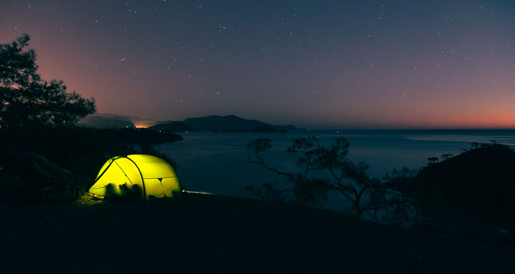 Tente Exped Venus II illuminée de nuit sur une falaise surplombant la mer Égée, bivouac sous un ciel constellé d'étoiles près de Fethiye, Turquie