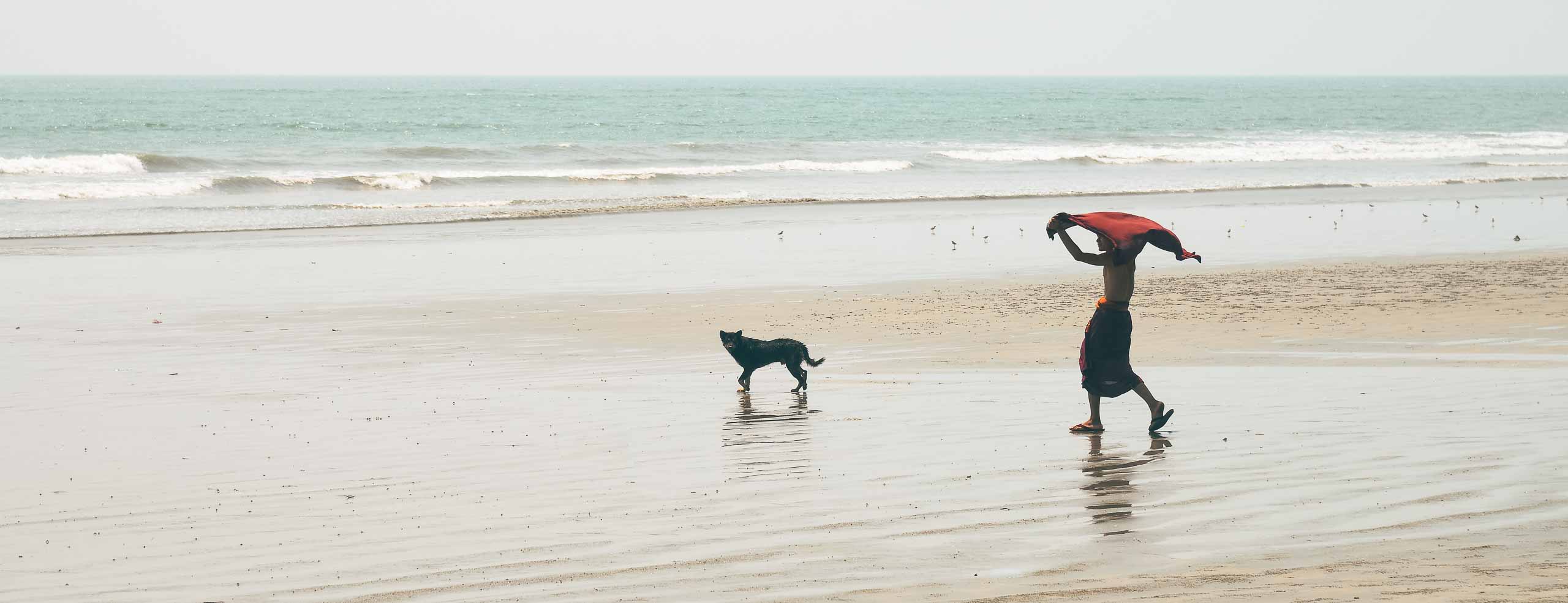Image épurée d'un jeune novice sur la plage de Maungmagan, il marche en retardant vers l'horizon, le kesa bouddhiste safran au vent, un chien à côté de lui