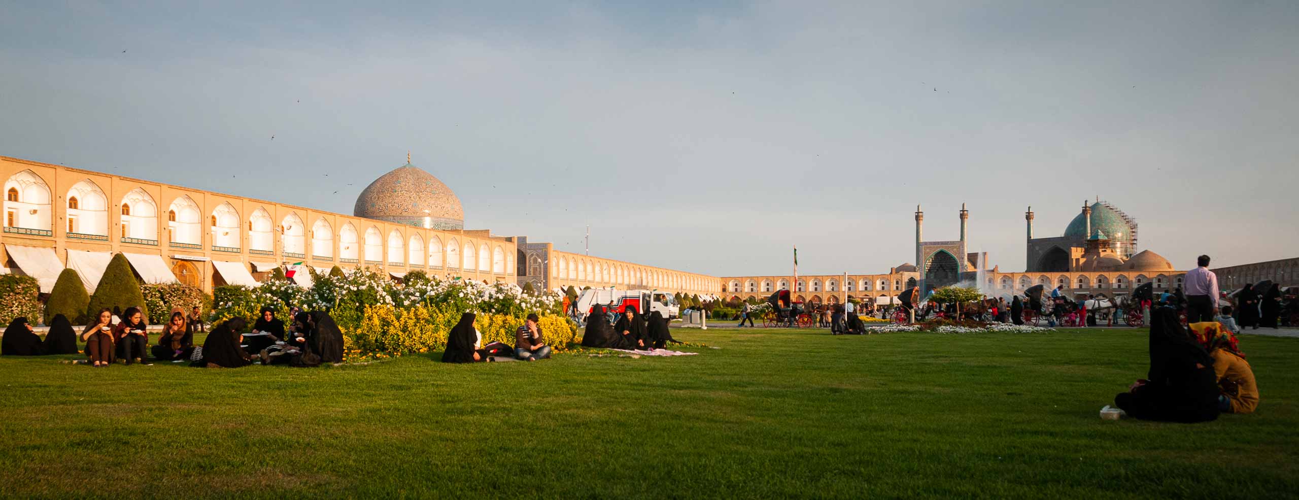 Photographie de la place Naqsh-e Jahan, des jeunes iraniens et iraniennes sont assis au soleil dans l'herbe, Meidan-e Emam, Isfahan, Iran