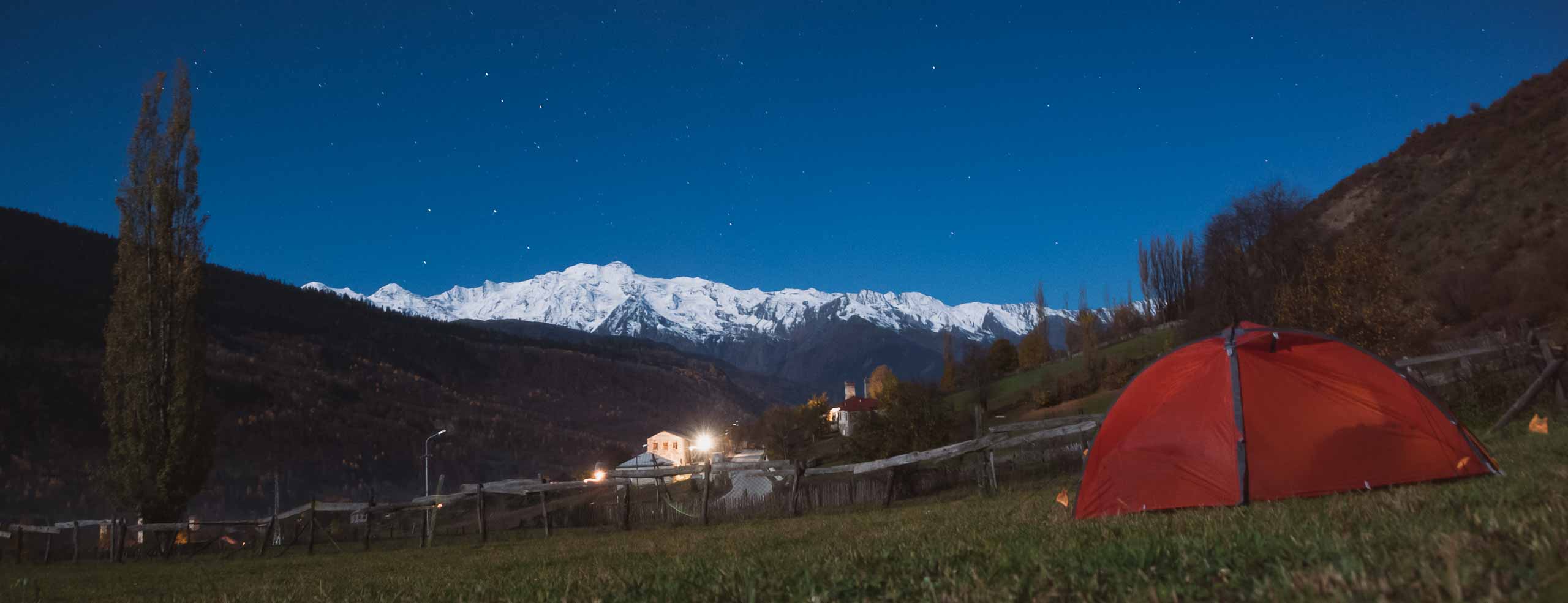 Bivouac dans le Haut-Caucase près de Mestia, Svaneti, Géorgie. Une tente rouge dans un champ avec des montagnes enneigée en fond, et un ciel étoilé