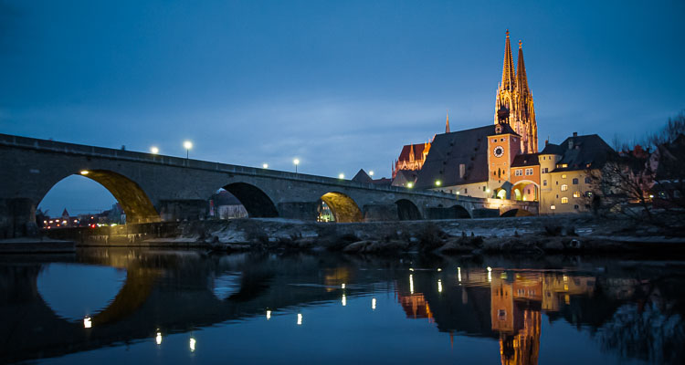 Rastibonne at night, a stone bridge crossing the Elbe in front of the Gothic cathedral of Regensburg, Germany. The reflection of the bridge and the cathedral is visible in the Elbe.