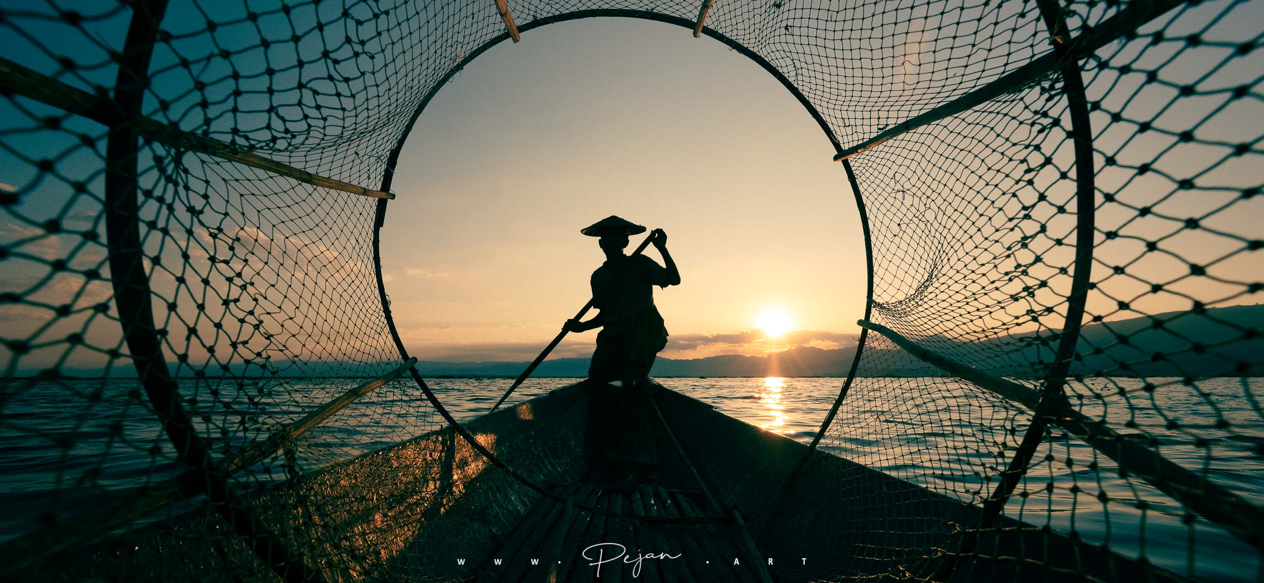 Silhouette d'un pêcheur Intha accroupi sur son bateau au lever du soleil. Photographie iconique prise à travers un filet circulaire, ce qui centre le sujet. Lac Inle, Birmanie.