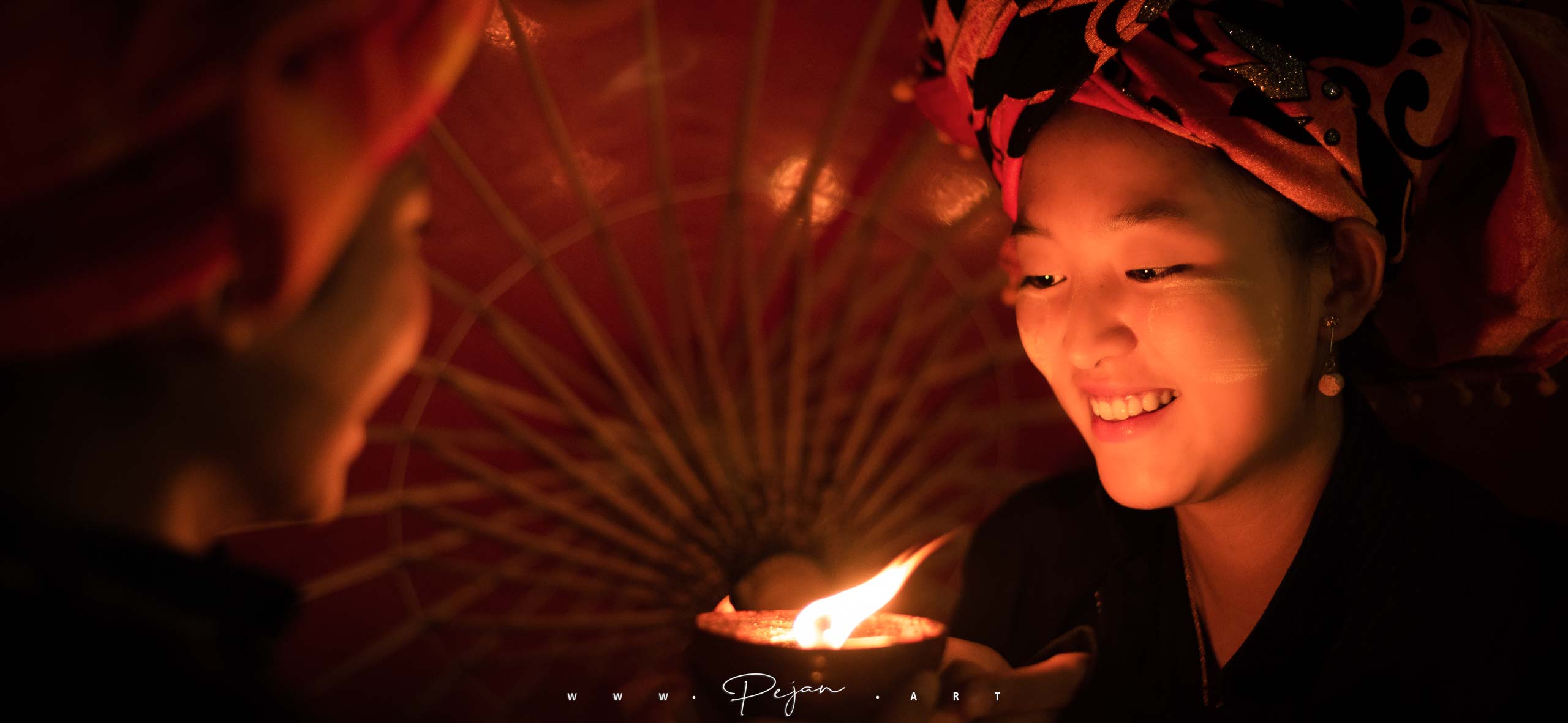 Portrait de nuit d'une jeune femme de l'ethnie Pao qui regarde en souriant les flamme d'une bougie devant une ombrelle. Elle porte du tanaka et une écharpe traditionnelle rouge de son ethnie. Lac Inle, Birmanie.