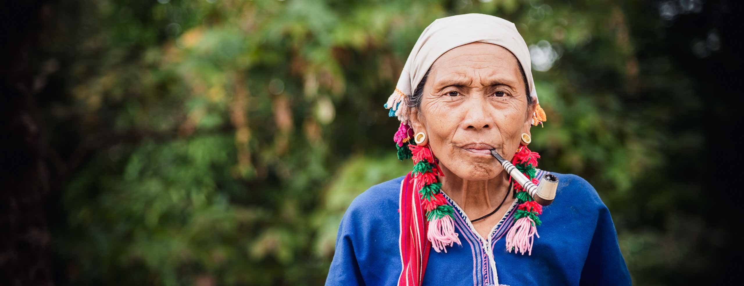 Portrait d'une femme de l'ethnie Karen rencontrée dans les montagnes en Thaïlande. Elle porte des vêtements et bijoux traditionnels et fume une grande pipe. Elle regarde l'objectif.